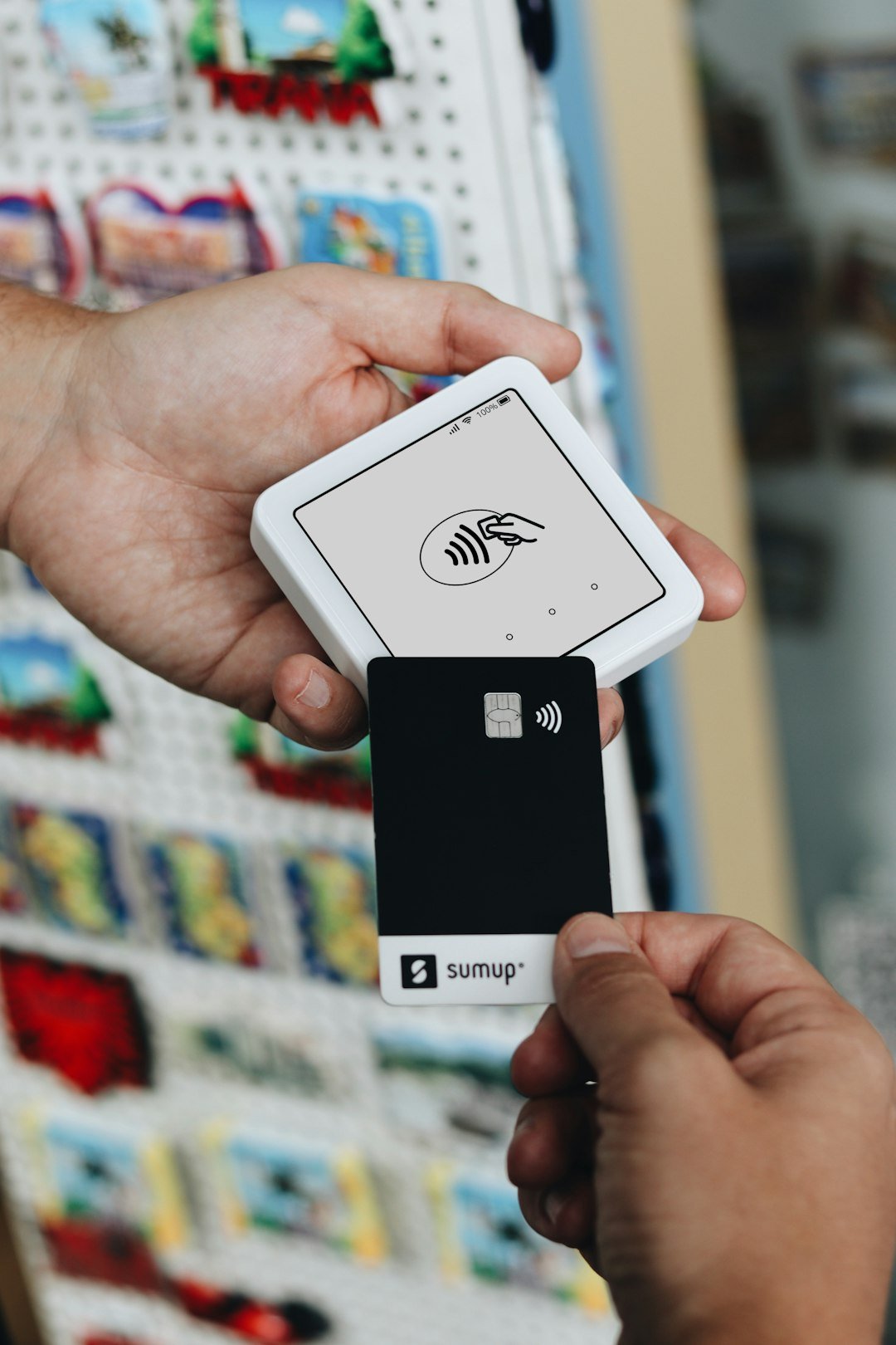 A customer is making a contactless payment using a SumUp card and a portable SumUp card reader. The card is being tapped on the reader, showing the seamless process of cashless payments. The background suggests a souvenir or retail shop environment, emphasizing the convenience and efficiency of using SumUps payment solutions for small businesses and merchants.